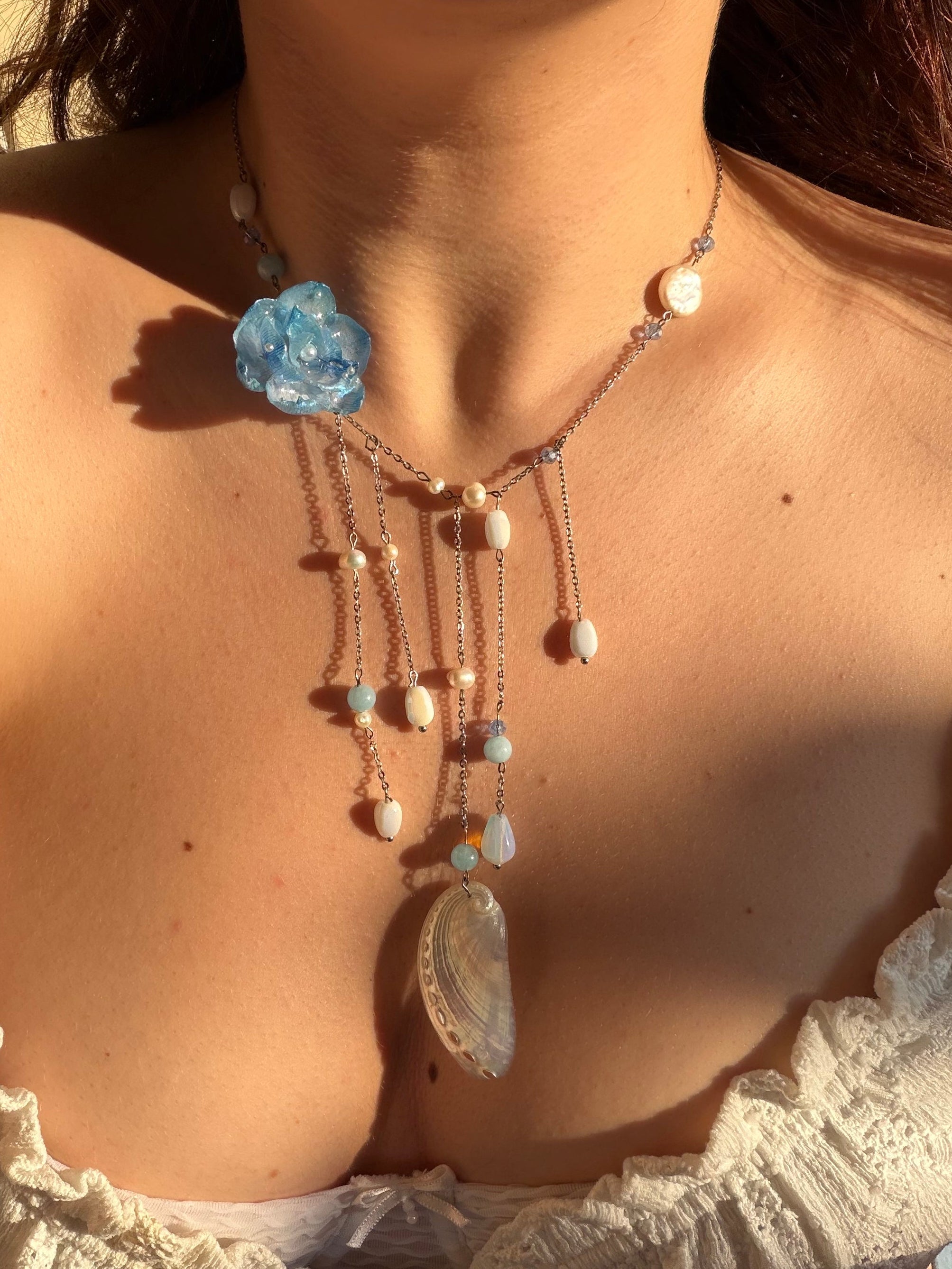 Necklace with blue flower pendant and pearl-like beads on a person wearing a white lace top.