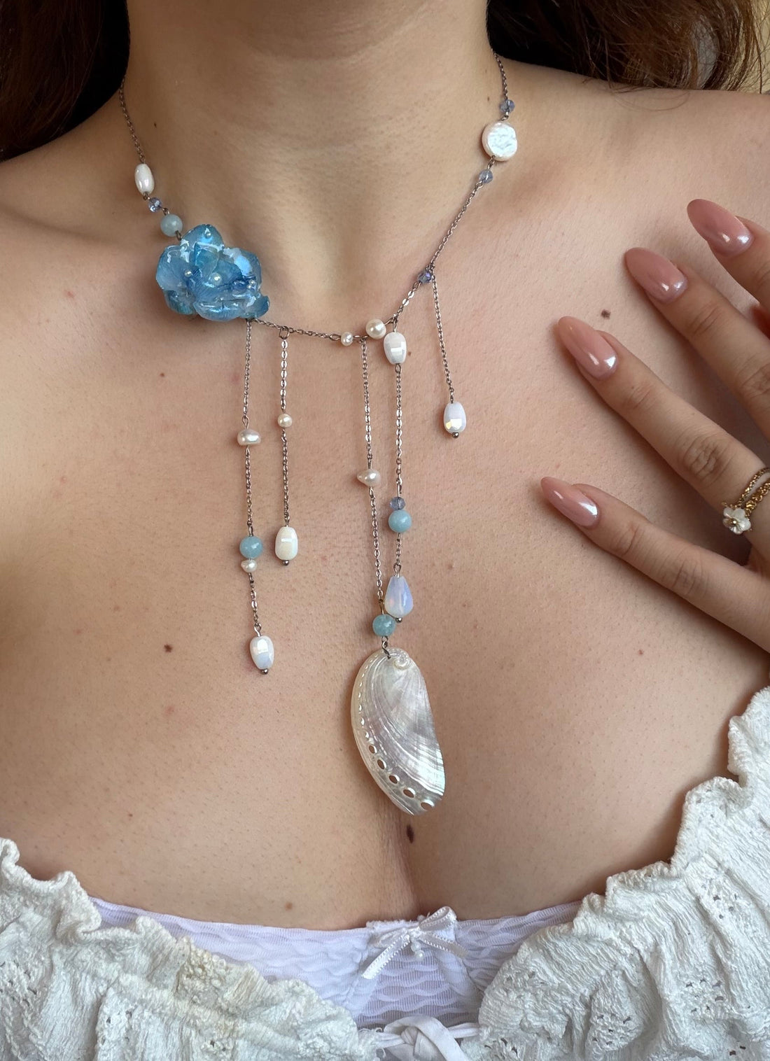 Necklace with blue flower pendant and pearl-like beads on a person wearing a white lace top.